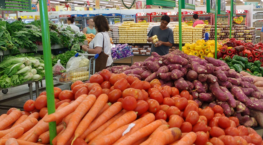 #PraCegoVer Frutas e legumes em primeiro plano, dispostos em gôndolas no supermercado