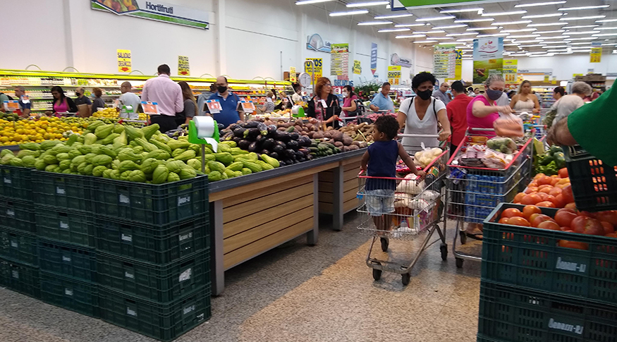 #PraCego Ver A foto mostra o interior de um supermercado, com pessoas empurrando carrinhos de compras no corredor.