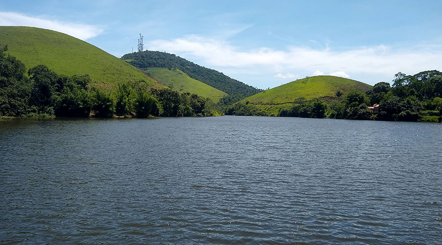#PraCegoVer foto de água de um açude no primeiro plano ao fundo montanhas verdes com torres de telefonia no topo