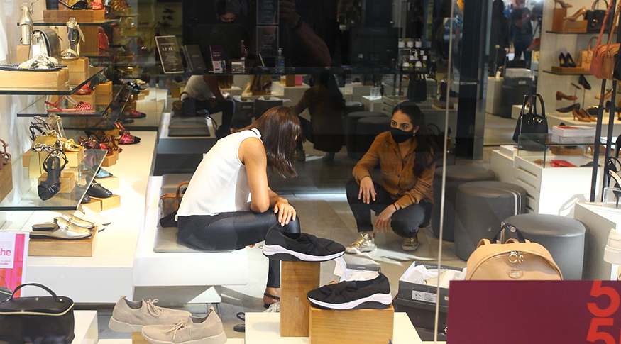 #PraCegoVer vendedora de sapato atendendo uma consumidora que experimenta um par de sapato. a vitrine exibe vários modelos de sapatos
