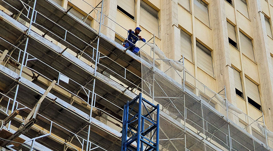 #PraCegoVer A foto mostra um operário de capacete, em cima de um andaime em uma obra de edifício