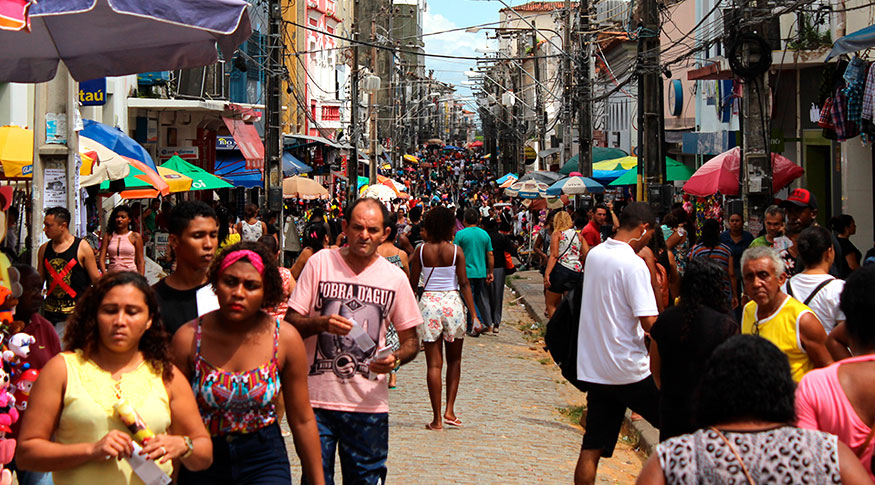 #PraCegoVer A foto mostra uma rua de comércio, com várias pessoas caminhando por ela