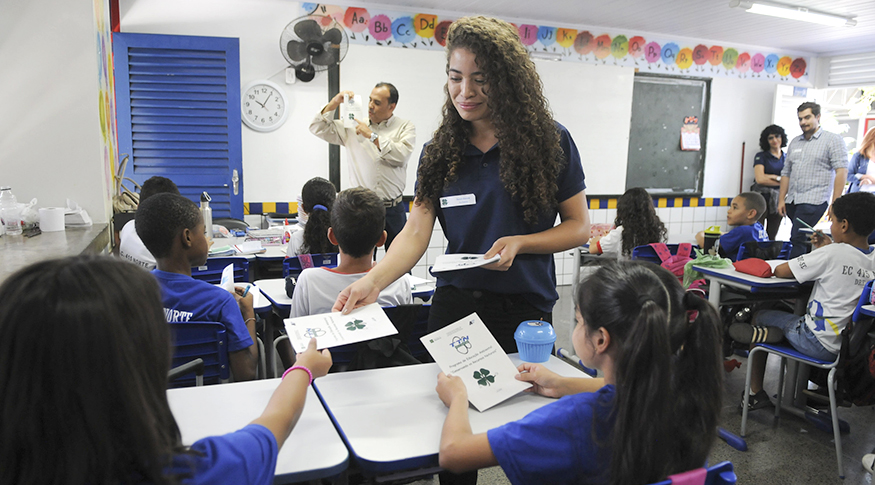 #PraCegoVer A foto mostra uma professora distribuindo material didático para sua turma de alunos, em sala de aula.