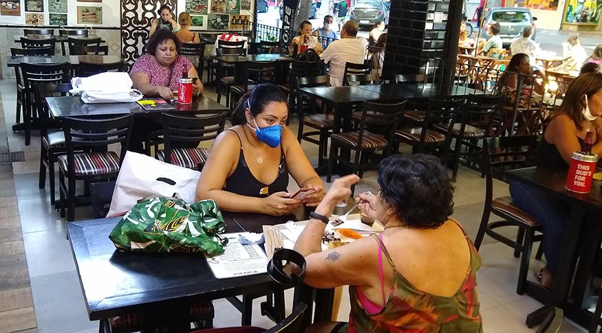 #PraTodosVerem A foto mostra pessoas sentadas em mesas de bar, algumas com máscaras.
