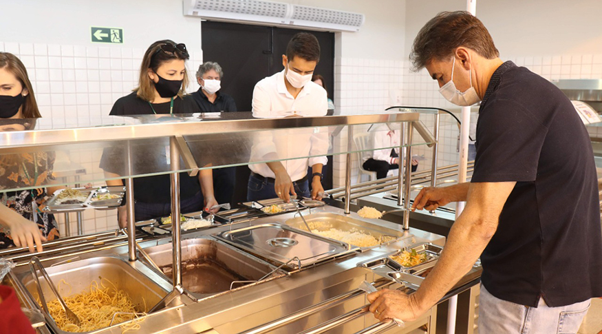 #PraTodosVerem A foto mostra um balcão de comidas de restaurante a quilo e clientes se servindo, todos usando máscara