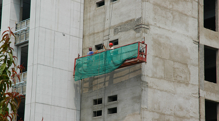 #PraCegoVer A foto mostra um andaime coberto com lona verde e dentro dele estão dois operários com capacetes. O andaime está preso num paredão cinza de um edifício em obras.
