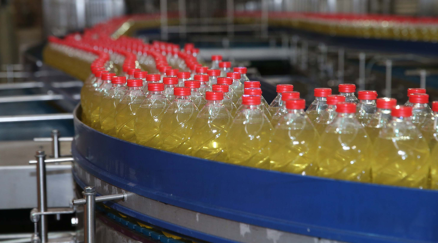 #PraCegoVer A foto mostra uma série de garrafas com líquido amarelo e tampas vermelhas, em linha de produção numa fábrica.