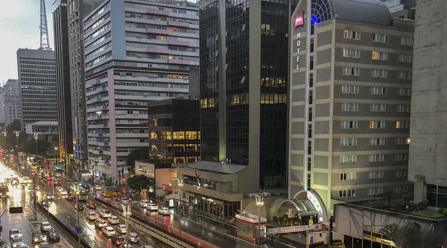 #PraCegoVer Vista aérea da cidade de SãoPaulo com a Avenida Paulista no canto inferior esquerdo da foto e 
 o hotel IBIS do lado dirito