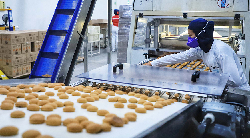 #PraCegoVer A foto mostra, do lado esquerdo, um funcionário de fáberica com EPIs e na sua frente uma bancada com bolinhos doces. Ao fundo, uma máquina industrial.