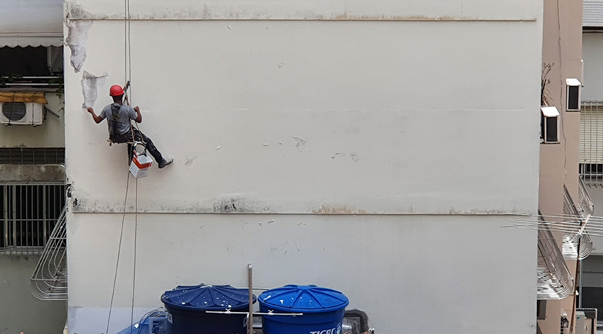 #PraCegoVer A foto mostra um operário de costas, com equipamentos de proteção, pendurado num andaime, pintando um paredão de um prédio.