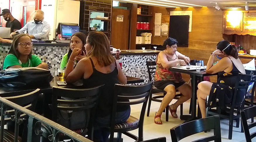 #PraCegoVer A foto mostra o interior de um restaurante, com mesas e cadeiras pretas. Na frente, do lado esquerdo, duas melhores estão sentadas conversando. Ao fundo, aparecem algumas pessoas sentadas comendo e conversando.