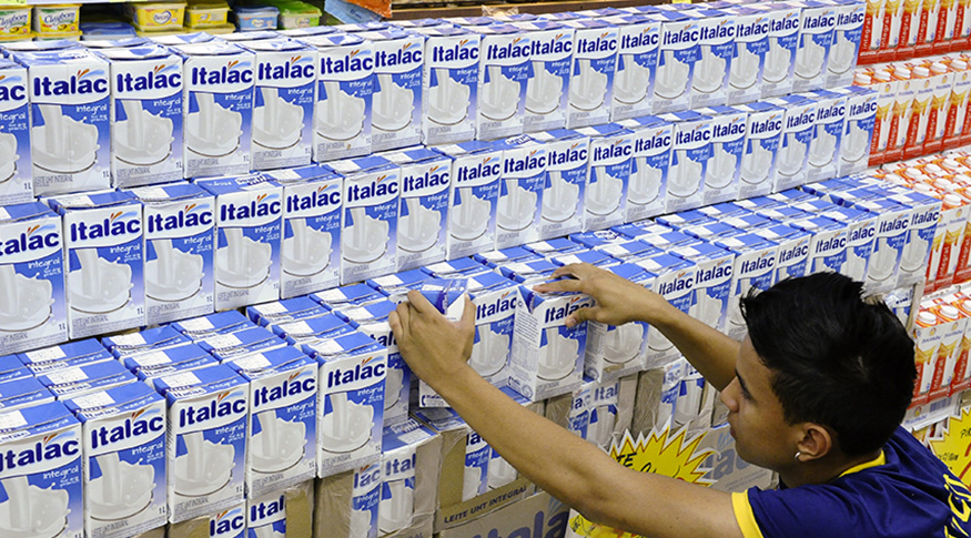 #PraCegoVer A foto mostra uma prateleira de supermercado lotada de caixas de leite longa vida e um empregado do mercado arrumando as caixas.