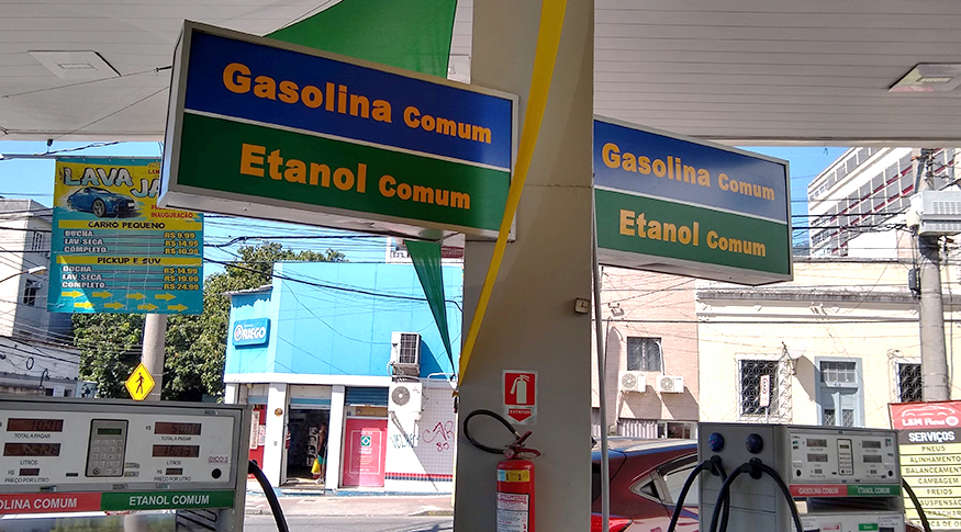 #PracegoVer A foto mostra um posto de gasolina com destaque para os letreiros que anunciam a gasolina comum, em azul e amarelo e o etanol comum, em verde e amarelo.