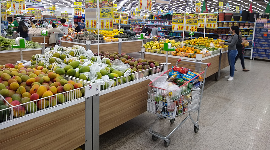 #PraCegoVer A foto mostra o stor de frutas dentro de um supermercado, com prateleiras cheias, carrinhos e pessoas ao fundo.