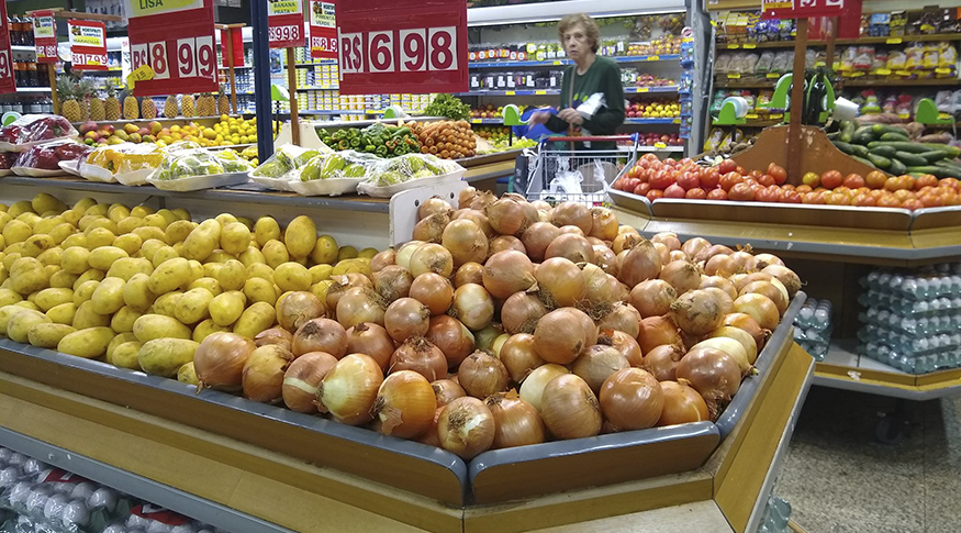 #PraCegoVer A foto mostra em primeiro plano, gôndolas de supermercado com cebola e batata.