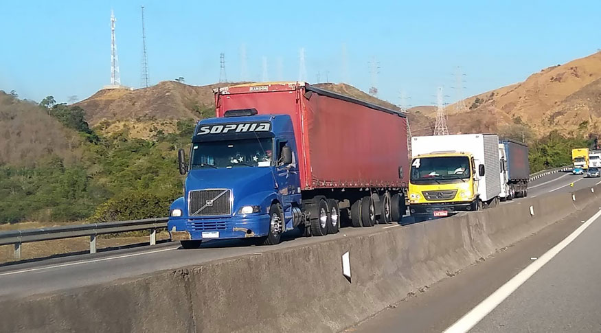 #PraCegoVer A foto mostra alguns caminhões de carga, sendo o primeiro com a caçamba vermelha, numa estrada larga.