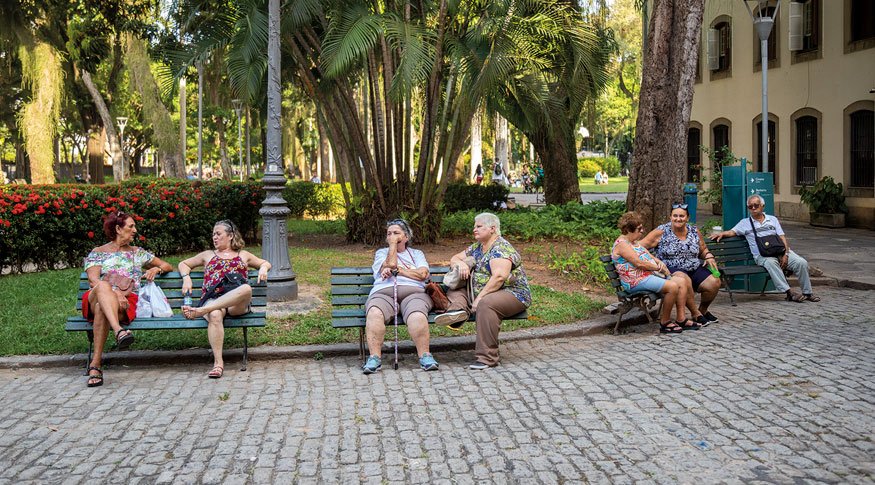 #PraCegoVer a foto mostra uma praça com idosos sentados em bancos de madeira