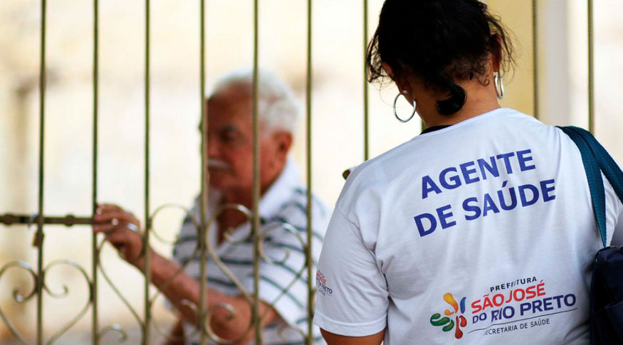 #PraCegoVer Mulher de costas vestida com camiseta branca,  uniforme de agente de saúde da prefeitura de São José do Rio Preto atende um senhor que está atrás de um portão de grade