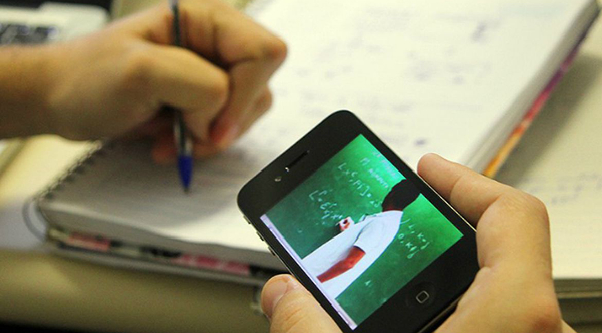 #PraCegoVer A foto mostra a mão de um aluno segurando um celular e na mesa um caderno de studos.
