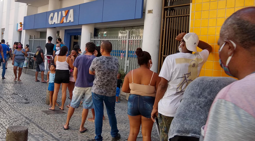 #PraCegoVer A foto mostra uma fila de pessoas na frente de um banco.