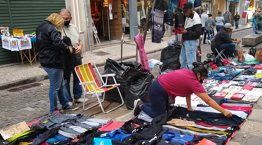 #PraCegoVer A foto mostra uma vendedora com seus produtos expostos na calçada de uma rua. Em volta, pedestres andam e olham sua mercadoria.