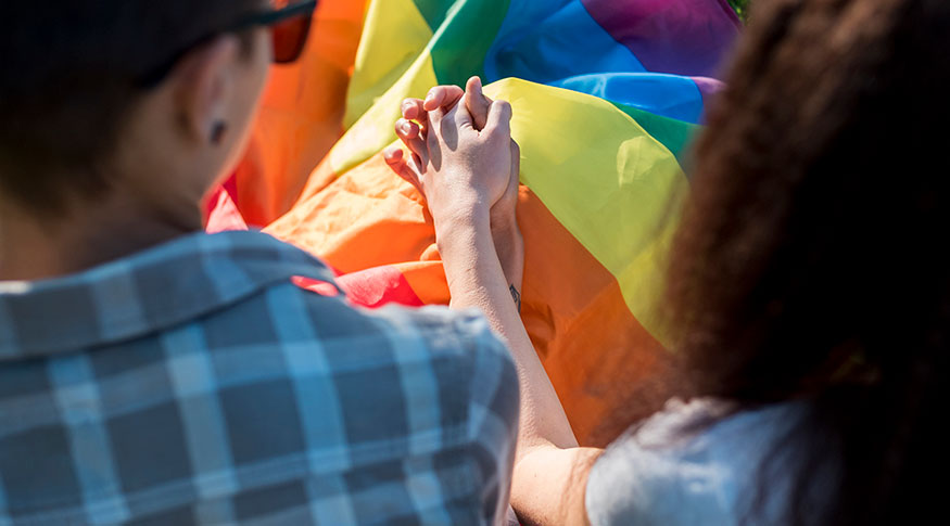 #PraCegoVer Duas mulheres de costas e de mãos dadas por cima da bandeira arco íris símbolo do movimento lgbtqia+