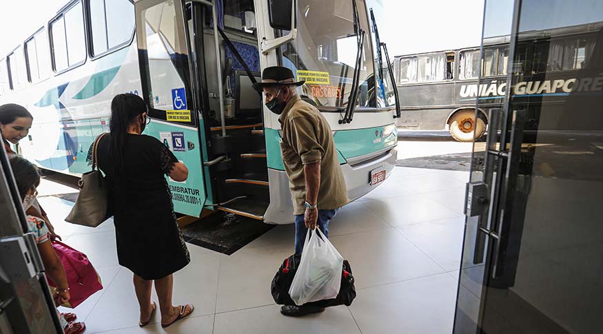 #PraCegoVer A foto mostra um ônibus de turismo ao fundo, 3 adultos e 1 criança se preparando para embarcar, numa rodoviária.