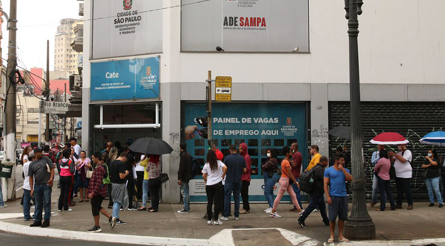 #PraCegoVer A imagem mostra uma fila de pessoas na rua perto de uma agência de empregos.