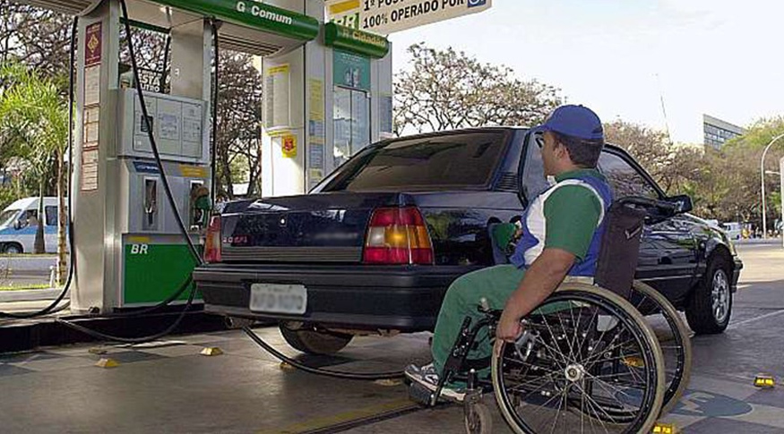 Em primeiro plano, homem preto em cadeira de rodas trabalha como frentista em um posto de gasolina, abastecendo um carro.  Ele veste uniforme com calça verde, camisa verde, branca e azul. Ao fundo, vê-se a bomba de gasolina que está sendo utilizada.