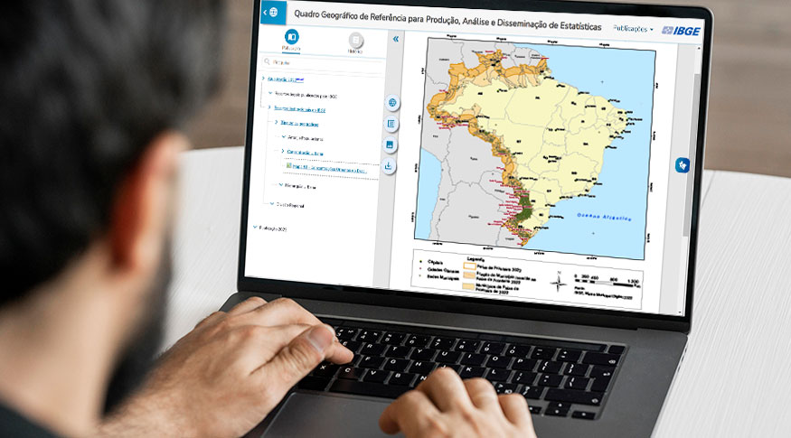 #PraCegoVer Homem de costas desfocado em primeiro plano usando laptop que está exibindo a tela da plataforma do Quadro Geográfico exibindo o mapa dos municípios do Brasil