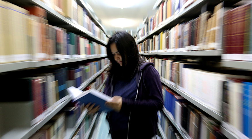 #PraCegoVer A foto mostra uma mulher ao centro, segurando um livro. No fundo, está a biblioteca, com efeito desfocado.
