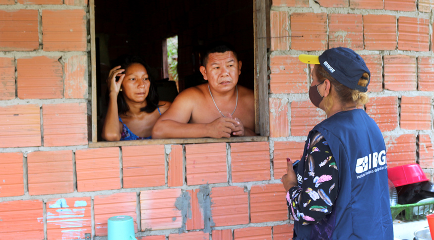 #PraCegoVer Recenseadora de costas entrevistando casal indígena pela janela da casa de tijolos