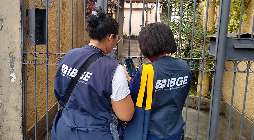 #PraCegoVer Duas mulheres recenseadoras vestindo uniforme do IBGE de costas em um portão de grade 