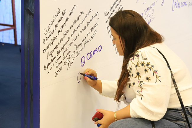 #PraCegoVer A foto mostra uma mulher escrevendo no mural do estande do IBGE.